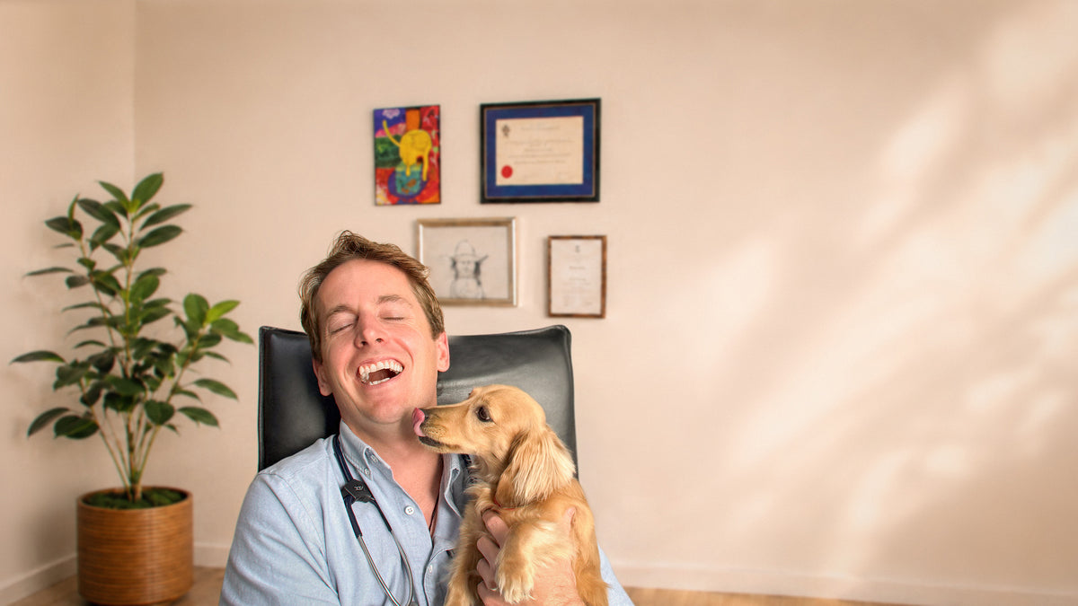 Veterinarian Dr. Feargus McConnell laughing as Frankie, a Dachshund, licks his face, highlighting the caring, trusted approach behind Dr. Ferg's dog health education and supplements.